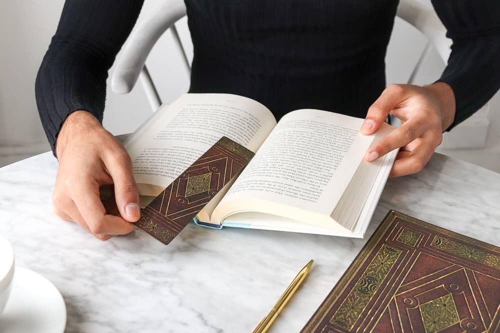 Person holding an open book on a marble surface with a pen and another book.