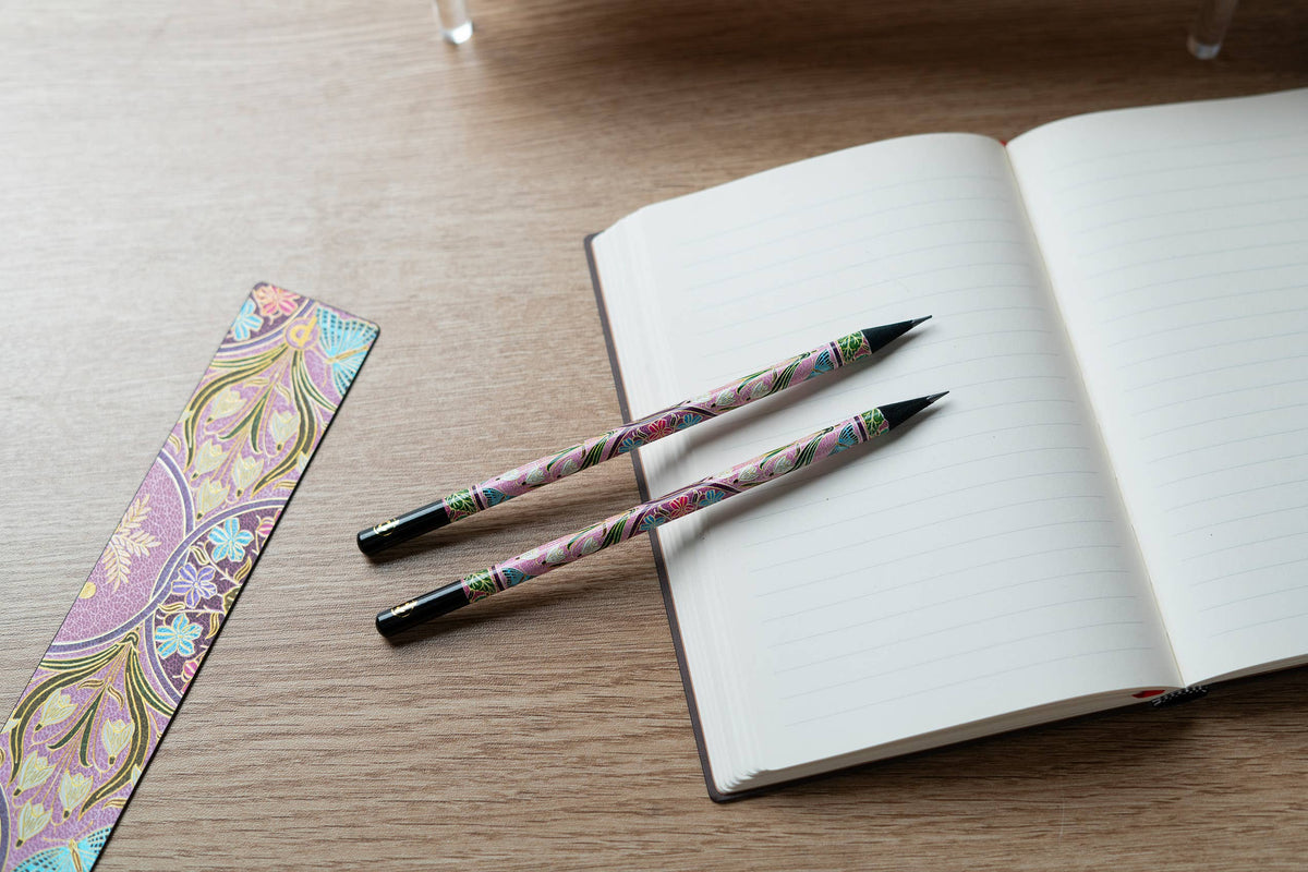 Two pens with colorful designs on a wooden surface next to an open notebook and a decorative bookmark.