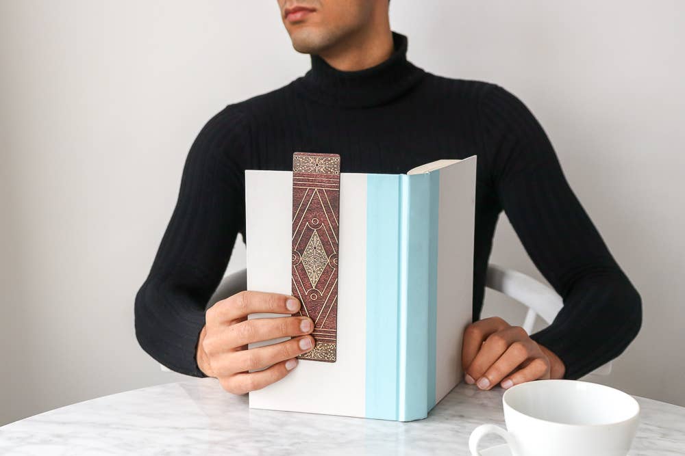 Person holding a book with a decorative cover on a table with a cup.
