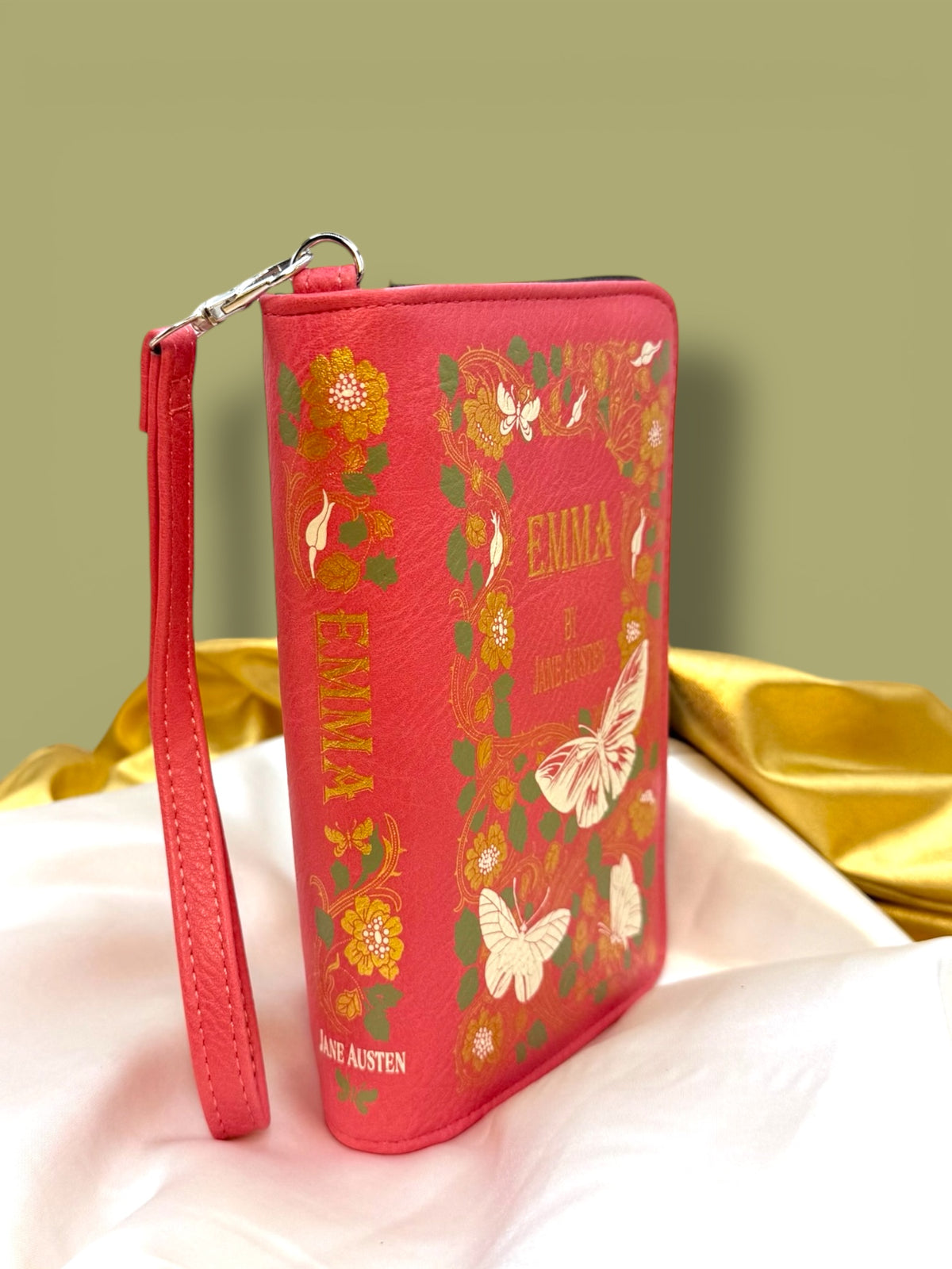 Emma Book Wallet inspired by Jane Austen, standing upright at an angle to show its pink and coral book-cover design, side profile, and wristlet strap.