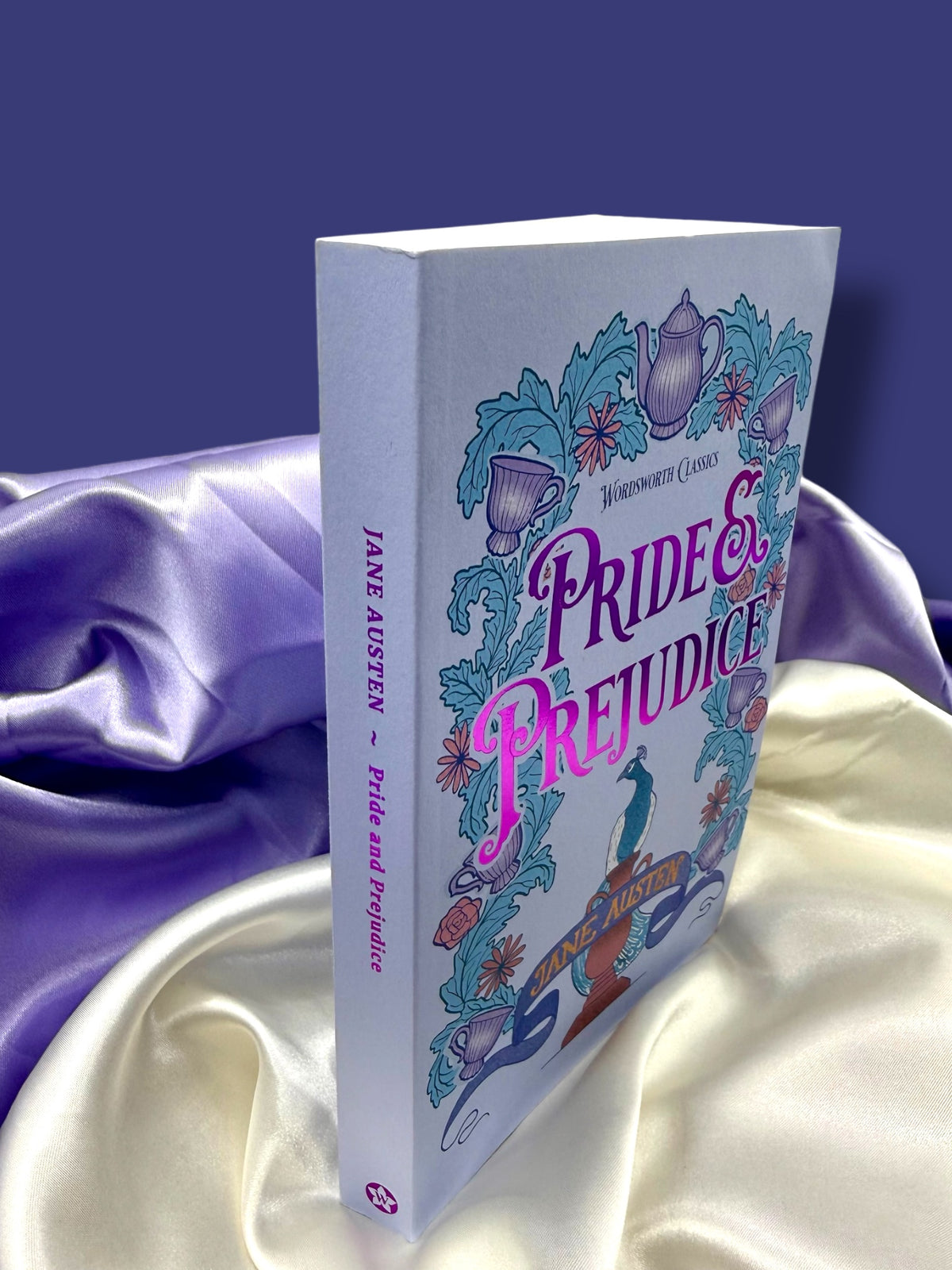 Angled view of Pride and Prejudice by Jane Austen, Wordsworth Classics paperback standing against purple satin fabric, showing its illustrated front cover and spine.