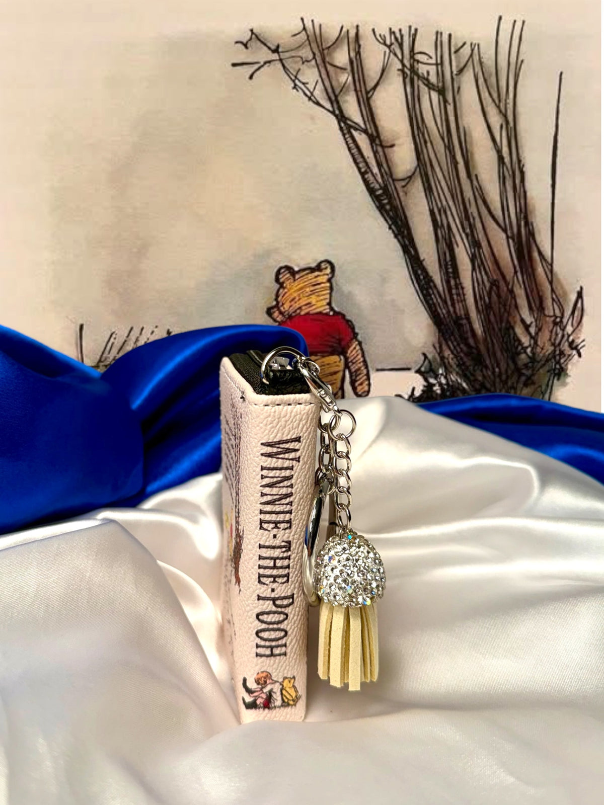 Side view of Winnie-the-Pooh book-shaped bag charm showing the illustrated spine, silver keychain, and cream tassel with sparkling cap.