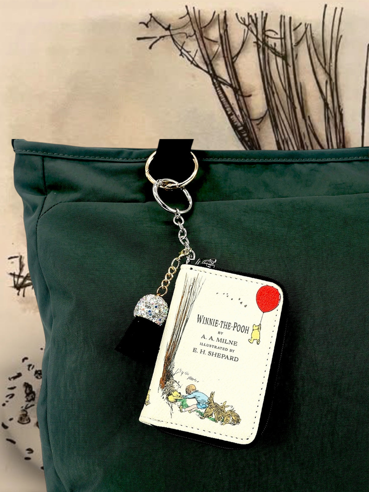 Winnie-the-Pooh miniature book bag charm hanging from a dark green handbag by its silver keyring, showing the illustrated cover and decorative tassel.