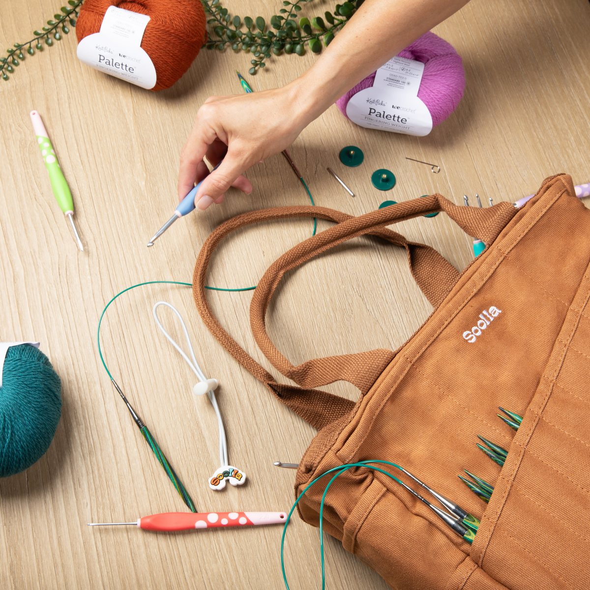 Person working with knitting needles and yarn on a wooden surface, with a brown Soolla Studio Bag containing needles.