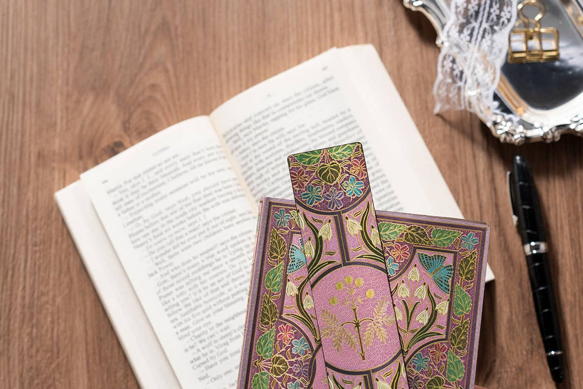 A decorative pink and gold pocket notebook with intricate floral patterns rests on top of an open book on a wooden desk, alongside an open book, a black pen, and a silver tray.