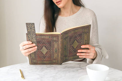 A woman in a beige knit sweater holds an open antique-style book with ornate brown and gold geometric cover detailing, sitting at a white marble table with a white cup nearby.