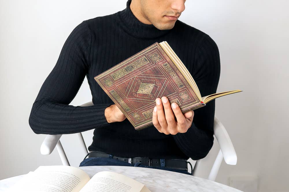 A man in a black ribbed turtleneck sweater sits in a white chair, holding an open antique-style book with a brown and gold geometric patterned cover. Another open book lies on a light marble table in the foreground against a plain light gray wall.