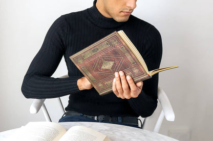A man in a black ribbed turtleneck sweater sits in a white chair, holding an open antique-style book with a brown and gold geometric patterned cover. Another open book lies on a light marble table in the foreground against a plain light gray wall.