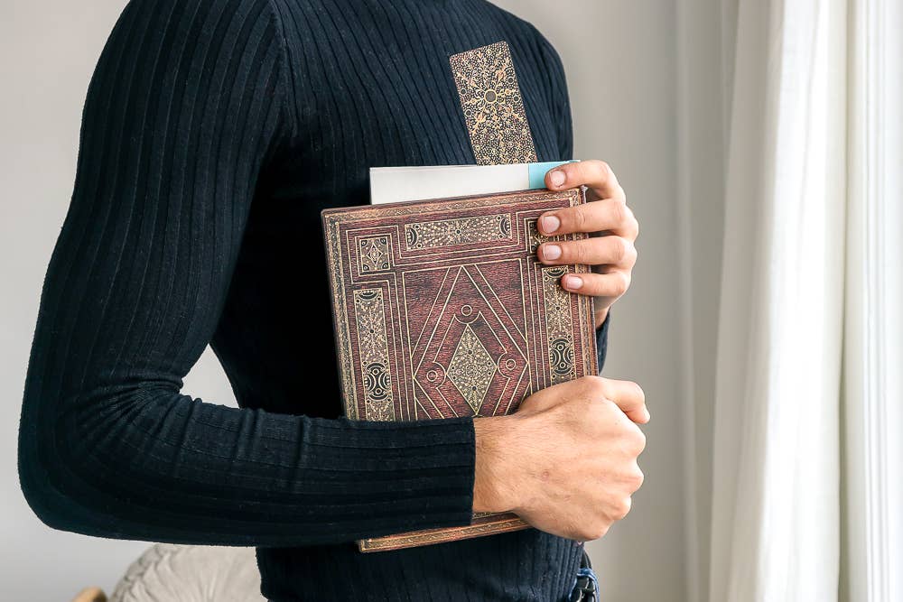 A person wearing a black ribbed long-sleeve top holds a brown faux-leather book or journal with ornate gold geometric patterns. The photo is taken from behind, showing the person&