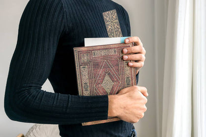 A person wearing a black ribbed long-sleeve top holds a brown faux-leather book or journal with ornate gold geometric patterns. The photo is taken from behind, showing the person&