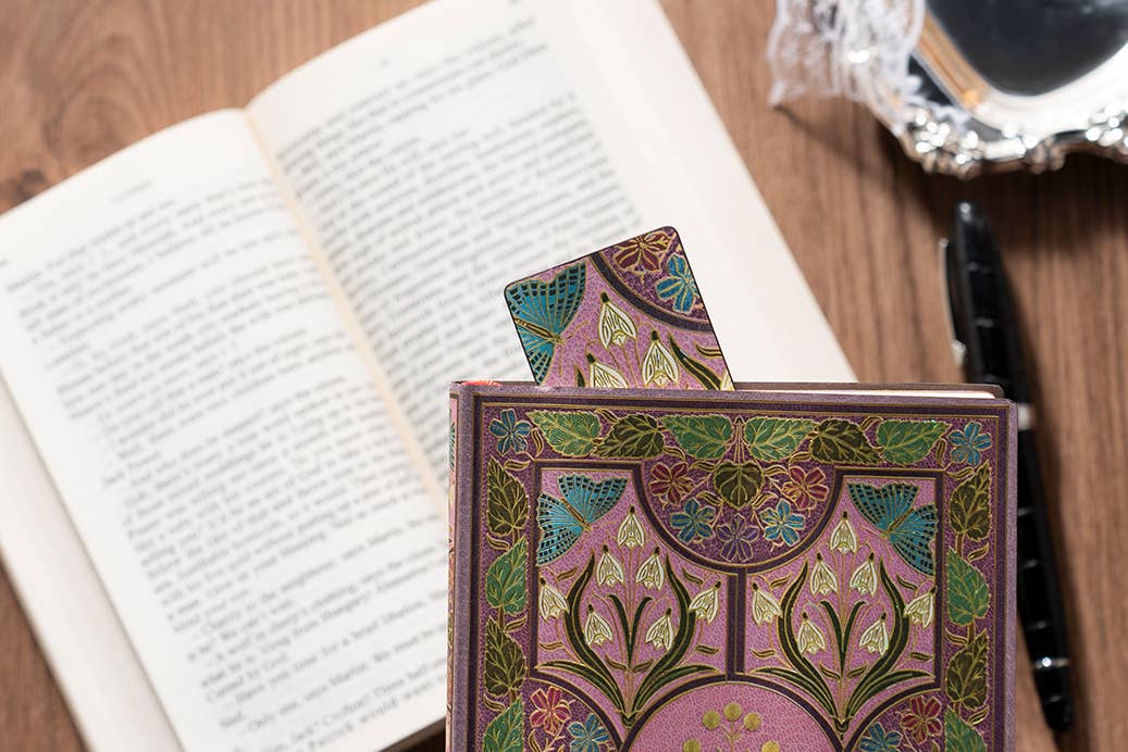 A decorative hardcover notebook or journal with an intricate Art Nouveau pattern in pink, green, and gold, standing on a wooden desk next to an open book.