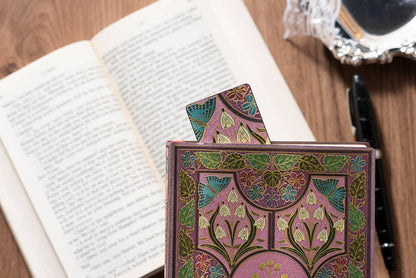 A decorative hardcover notebook or journal with an intricate Art Nouveau pattern in pink, green, and gold, standing on a wooden desk next to an open book.