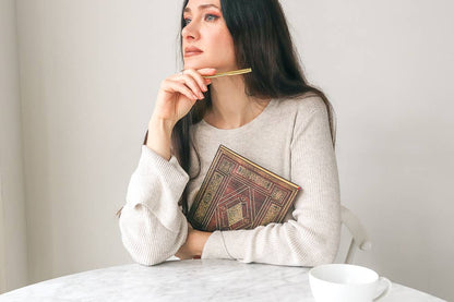 A woman with long dark hair sits at a table, holding a gold pen to her chin and resting a patterned book under her arm. She is wearing a light beige ribbed knit sweater, with a white mug on the marble table in front of her against a neutral light gray wall.