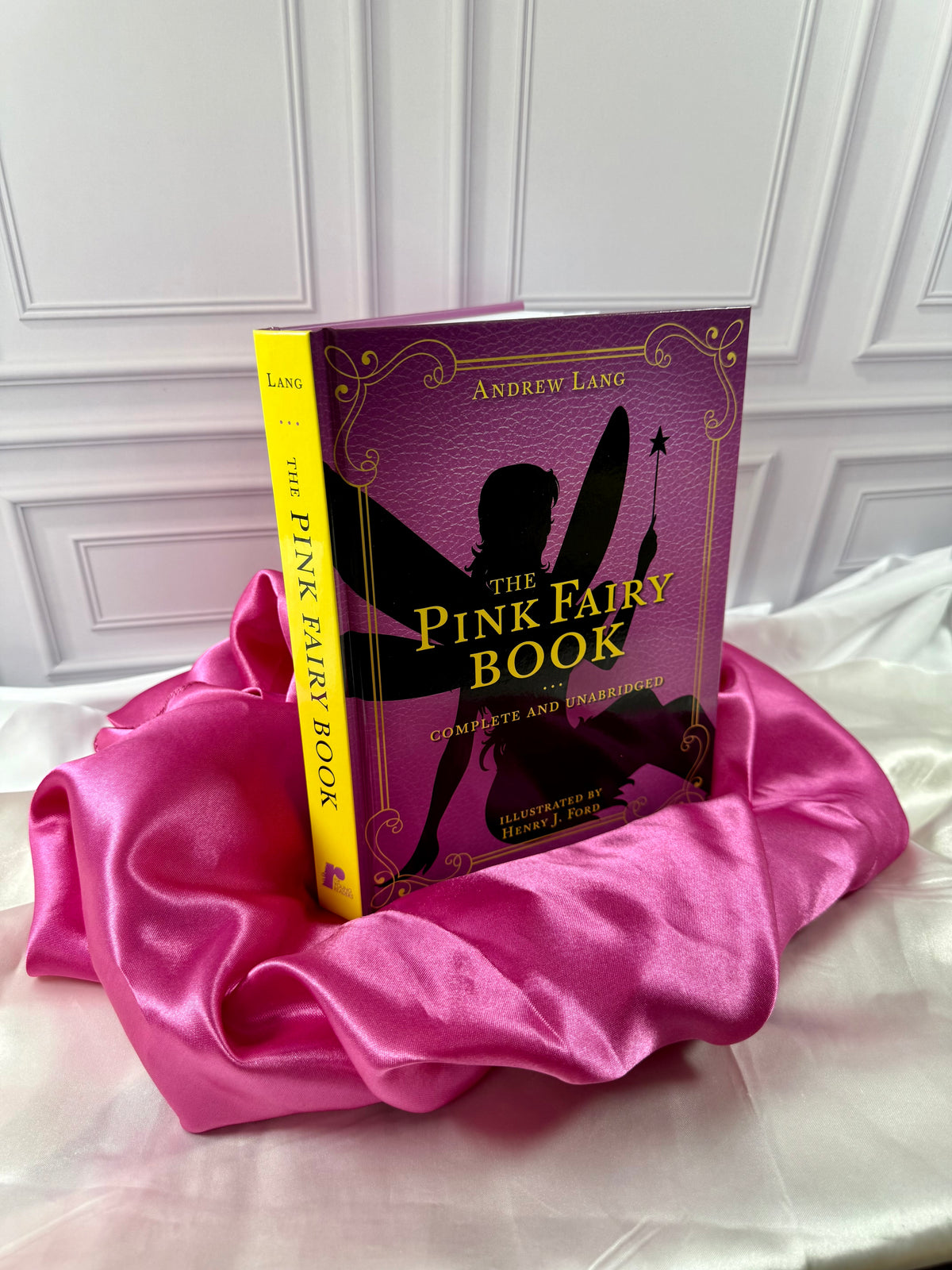 The Pink Fairy Book by Andrew Lang photographed at an angle, showing its bright yellow spine and decorative pink hardcover.