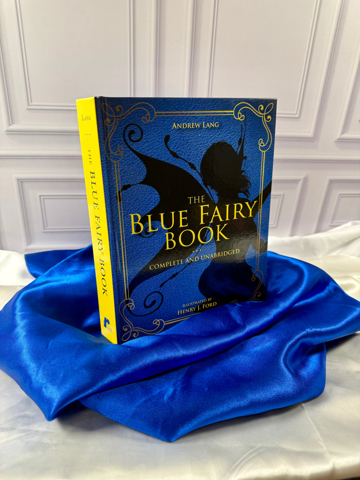 The Blue Fairy Book by Andrew Lang photographed at an angle, showing its bright yellow spine and decorative blue hardcover.
