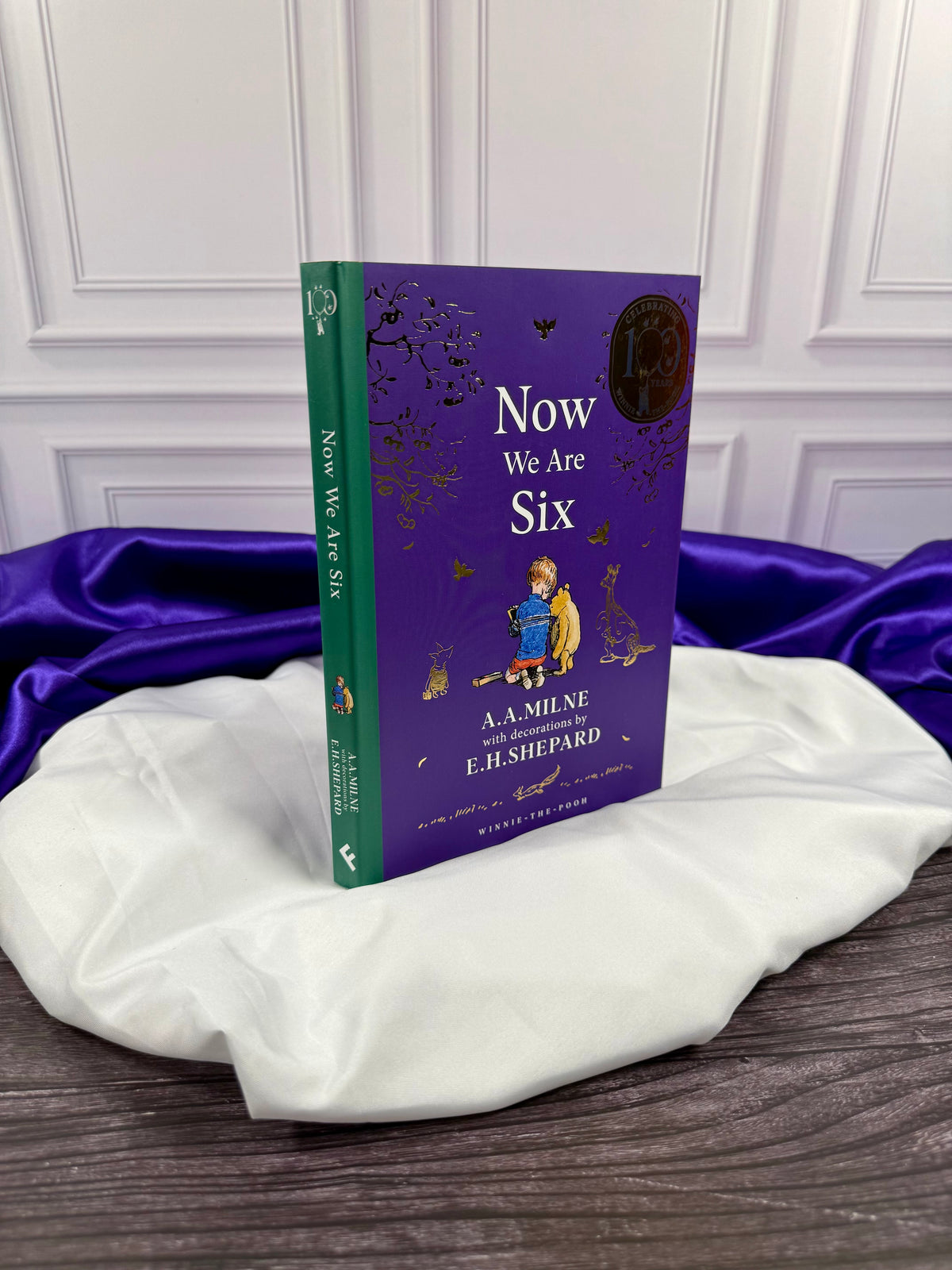 Now We Are Six illustrated hardcover by A. A. Milne shown at an angle, displaying the purple front cover and green spine with white title lettering.