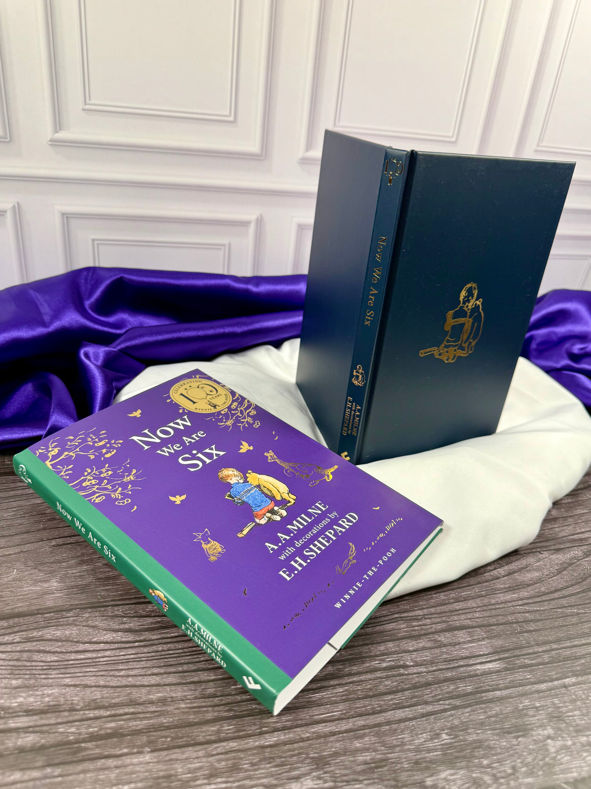 Now We Are Six by A. A. Milne showing the removable purple dust jacket beside the dark blue hardcover binding with gold lettering and Christopher Robin illustration.