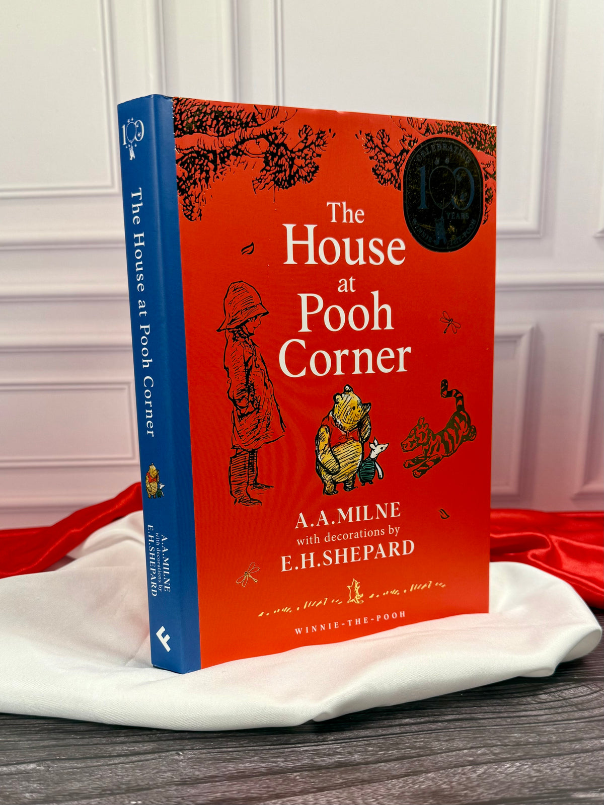 Angled view of The House at Pooh Corner anniversary hardcover showing its blue spine, orange-red illustrated dust jacket and E. H. Shepard artwork