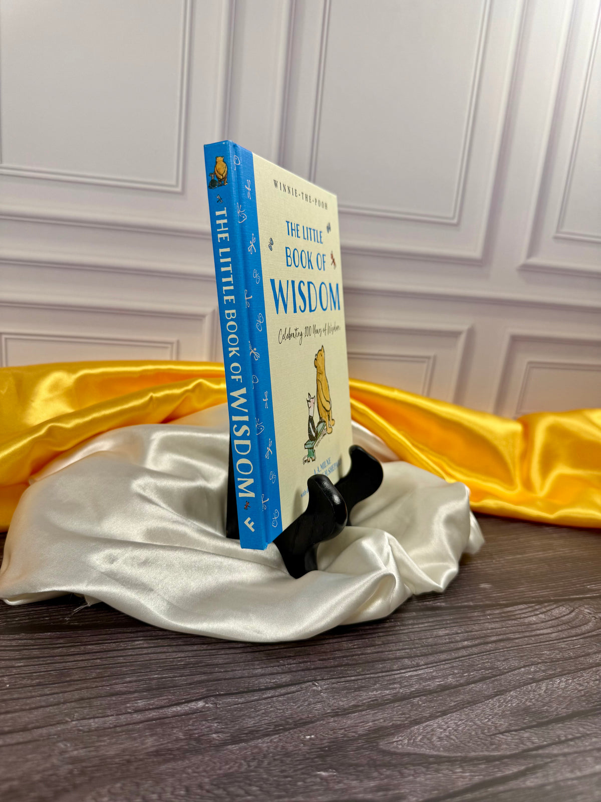 Winnie-the-Pooh: The Little Book of Wisdom hardcover by A. A. Milne shown at an angle, displaying its blue spine and illustrated yellow cover on satin fabric.