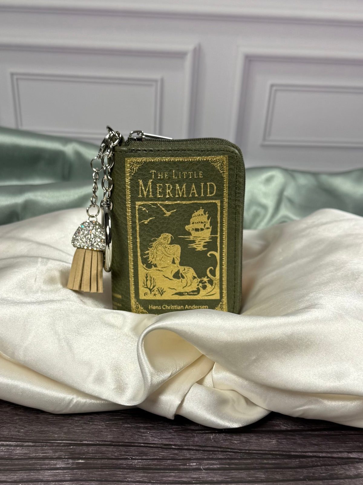 The Little Mermaid miniature book bag charm and coin purse with olive green cover, gold mermaid illustration, silver keyring, and decorative tassel.