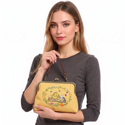 A woman holding the Winnie-the-Pooh and Piglet embroidered kisslock handbag featuring the two friends lounging on a log, linen blend fabric, and vintage-style metal clasp.