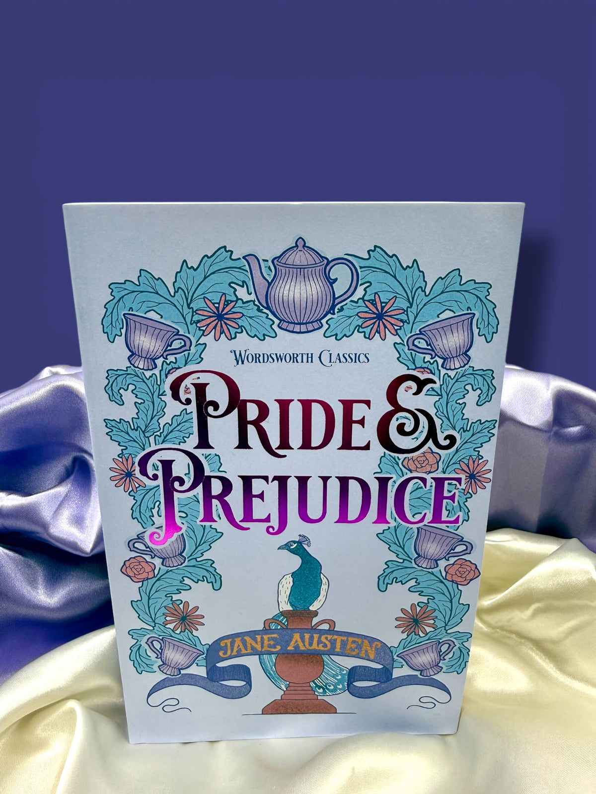 Pride and Prejudice by Jane Austen, Wordsworth Classics paperback with a colorful illustrated cover in blue, turquoise, and purple, displayed on satin fabric.
