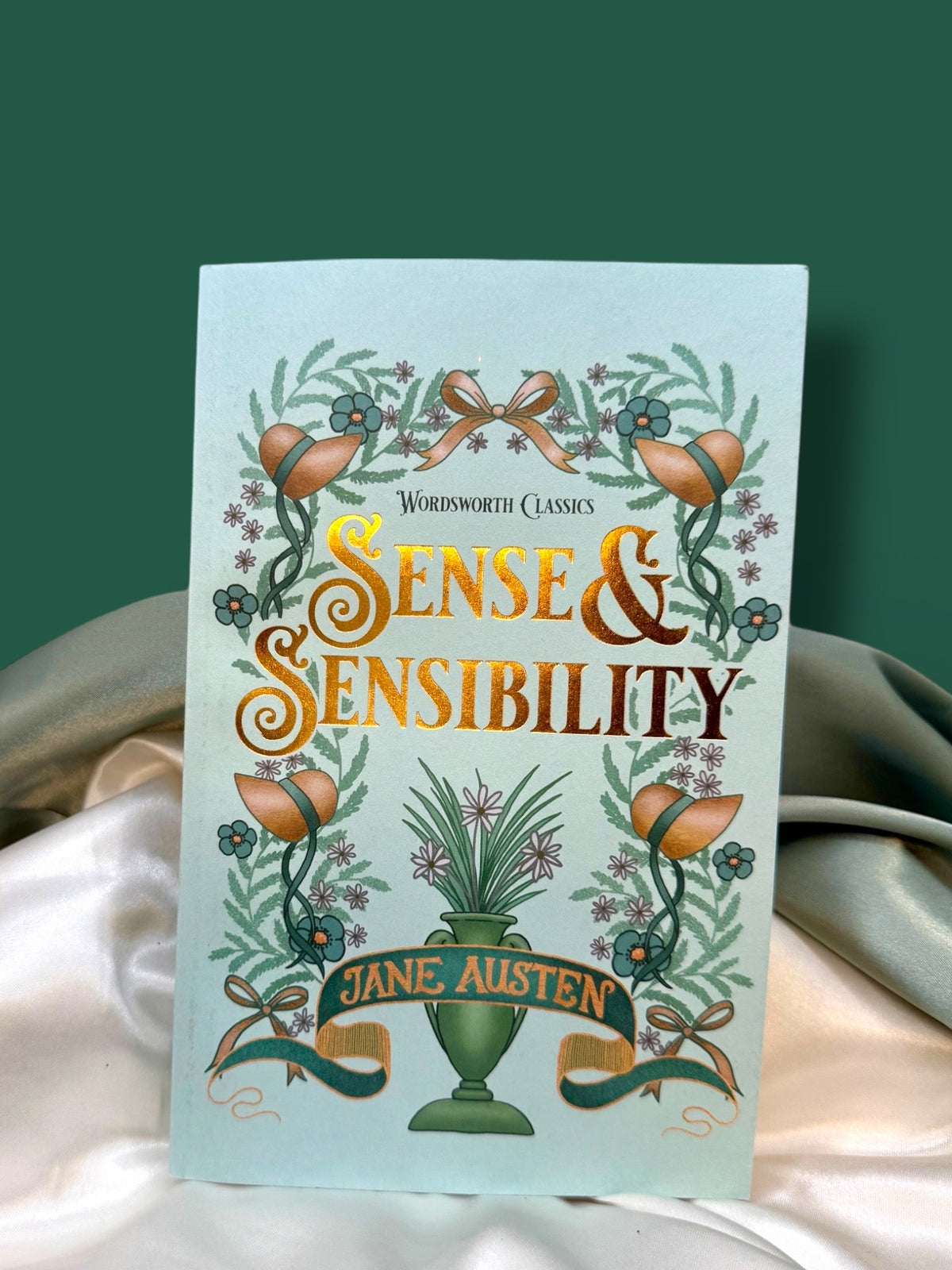 Sense and Sensibility by Jane Austen, Wordsworth Classics paperback with a pale mint-green cover, orange lettering, and decorative botanical illustration, displayed on ivory satin.
