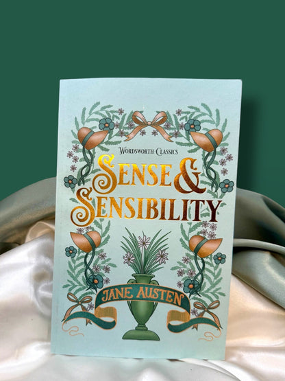 Sense and Sensibility by Jane Austen, Wordsworth Classics paperback with a pale mint-green cover, orange lettering, and decorative botanical illustration, displayed on ivory satin.