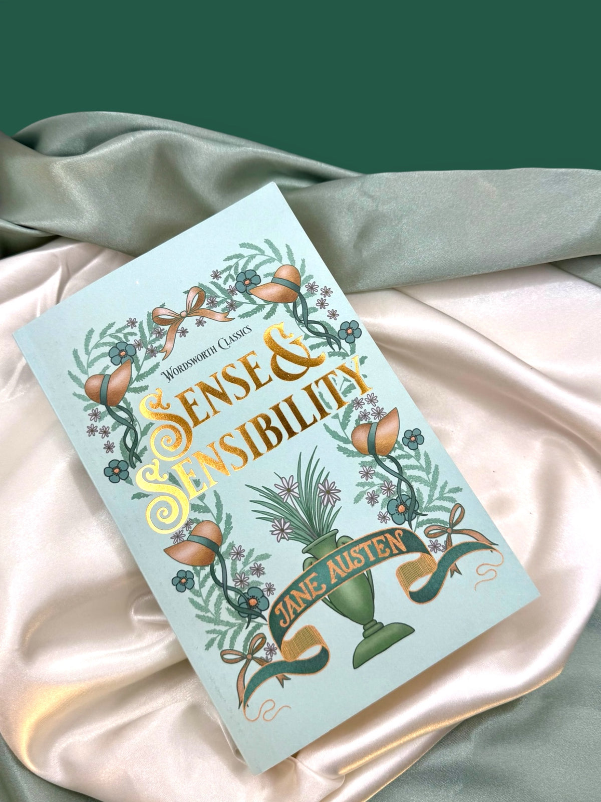 Sense and Sensibility by Jane Austen paperback resting on ivory satin, highlighting its mint-green cover and decorative botanical artwork.