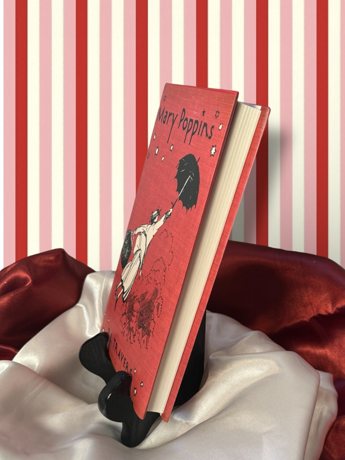 Side page edge and front view of the Mary Poppins hardcover edition by P. L. Travers, displayed on a book stand with red satin fabric.