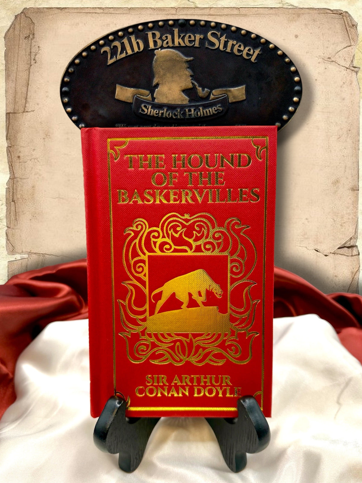 The Hound of the Baskervilles by Sir Arthur Conan Doyle, red clothbound hardcover with gold-embossed hound illustration and ornate decoration, displayed beneath a 221B Baker Street sign.
