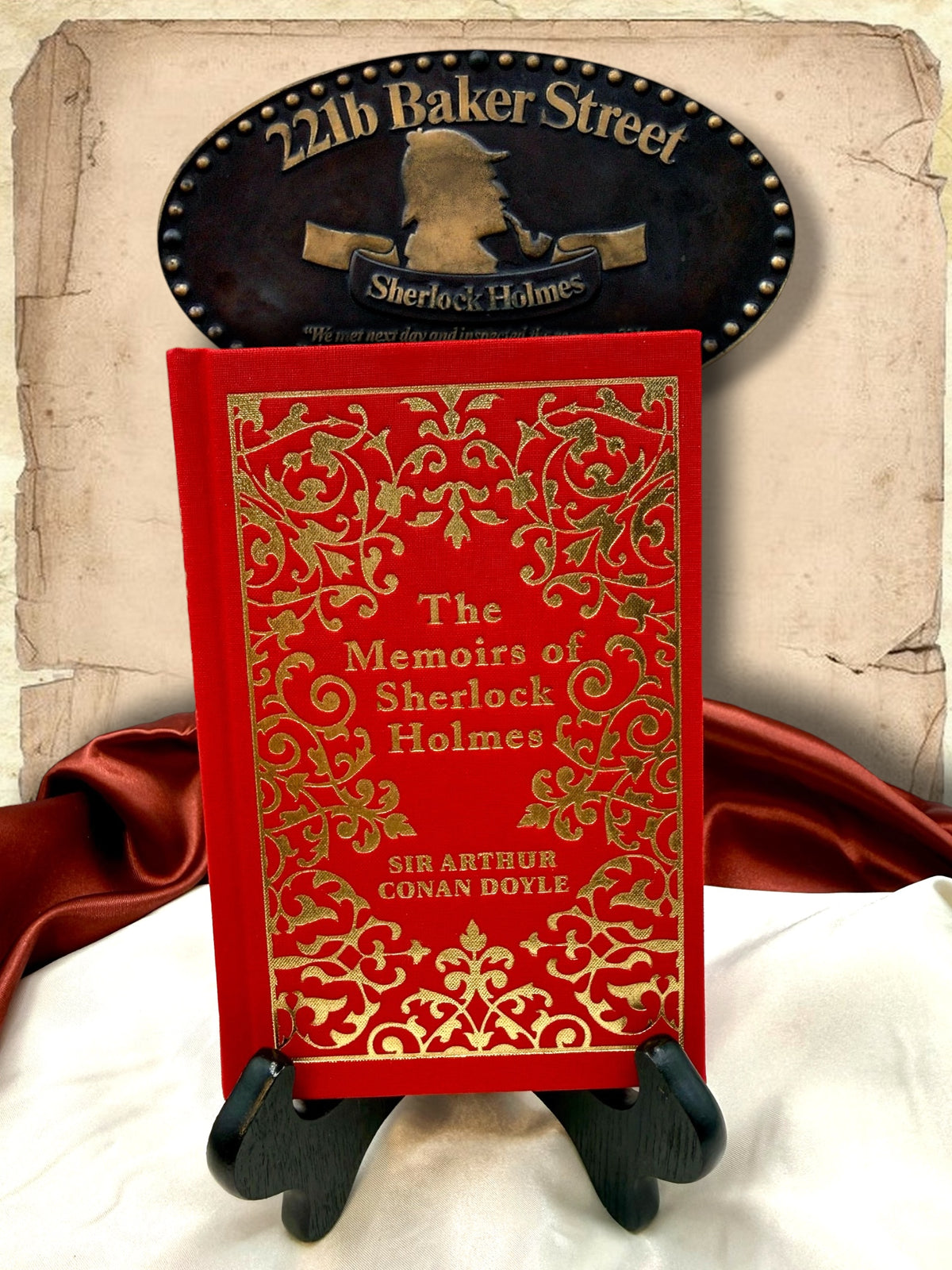 The Memoirs of Sherlock Holmes by Sir Arthur Conan Doyle, red clothbound hardcover with elaborate gold floral ornamentation, displayed on a wooden book stand beneath a 221B Baker Street sign.