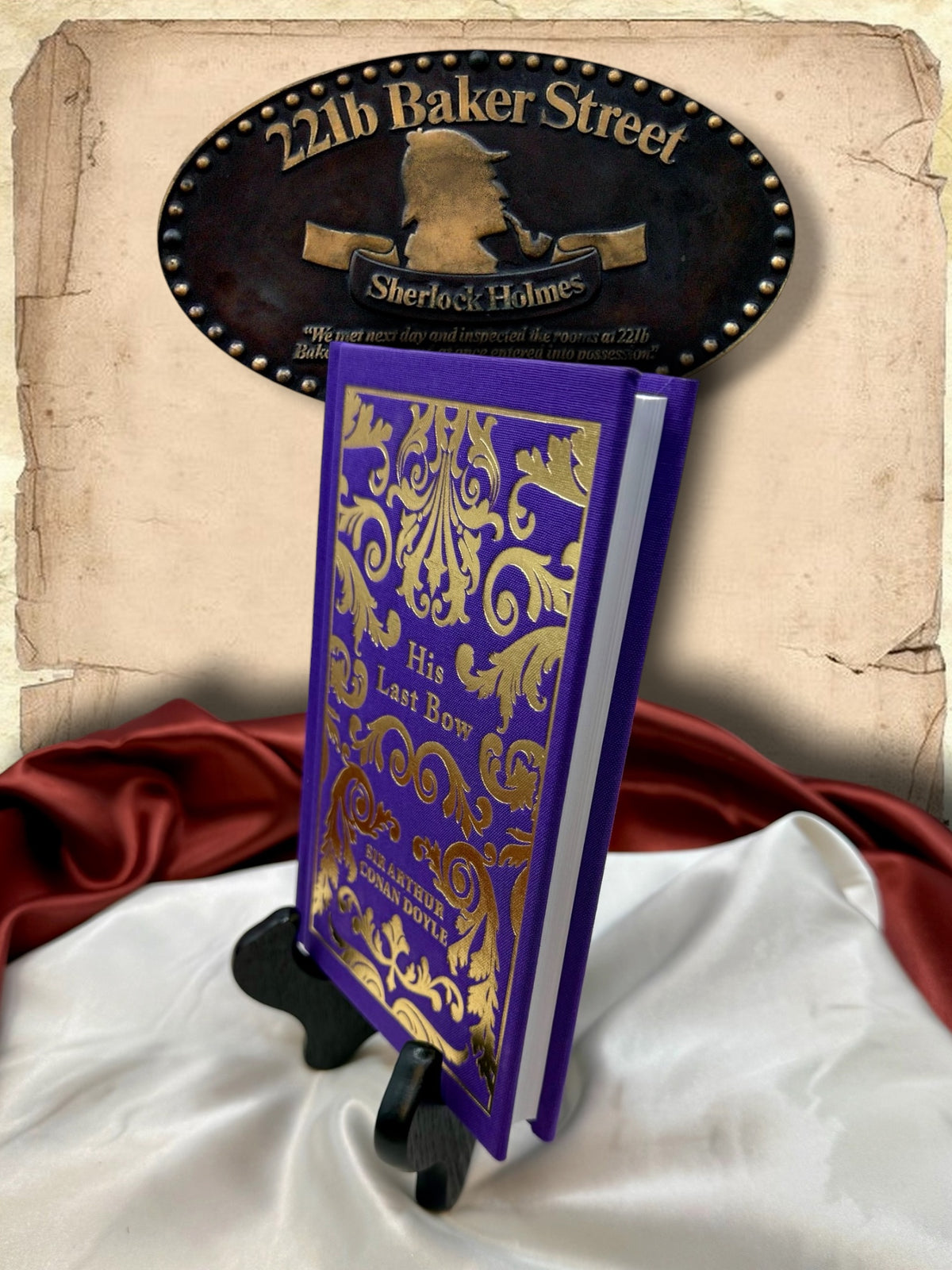 Three-quarter view of His Last Bow hardcover showing its purple cloth binding, ornate gold cover design, and white page edges on a wooden display stand.