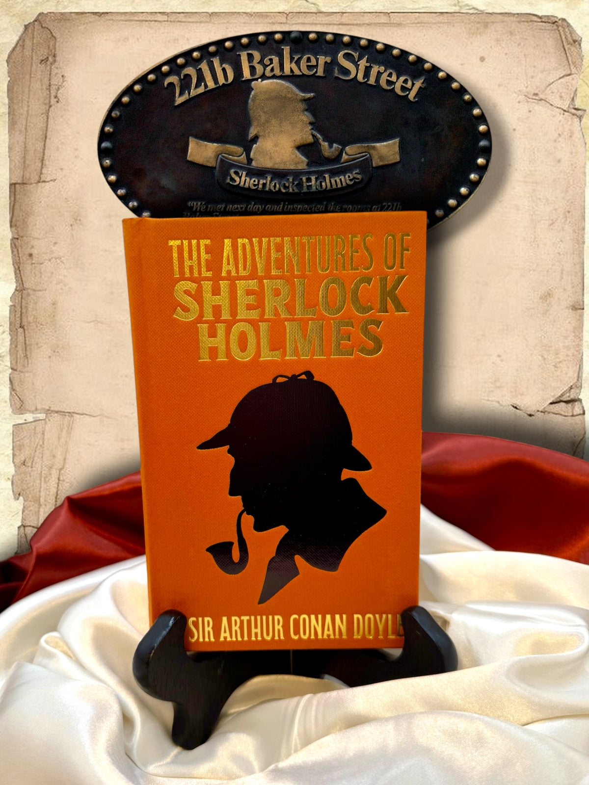 Orange hardcover The Adventures of Sherlock Holmes by Arthur Conan Doyle displayed beside a red Sherlock Holmes book wallet, with a 221B Baker Street plaque and satin fabric.