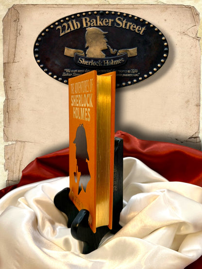 Side view of The Adventures of Sherlock Holmes orange hardcover displaying its reflective gold-gilded page edges, decorative cover, and black book stand.