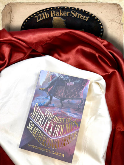 The Best of Sherlock Holmes Wordsworth Classics paperback lying on ivory satin fabric with burgundy draping, displaying the illustrated front cover and gold lettering.