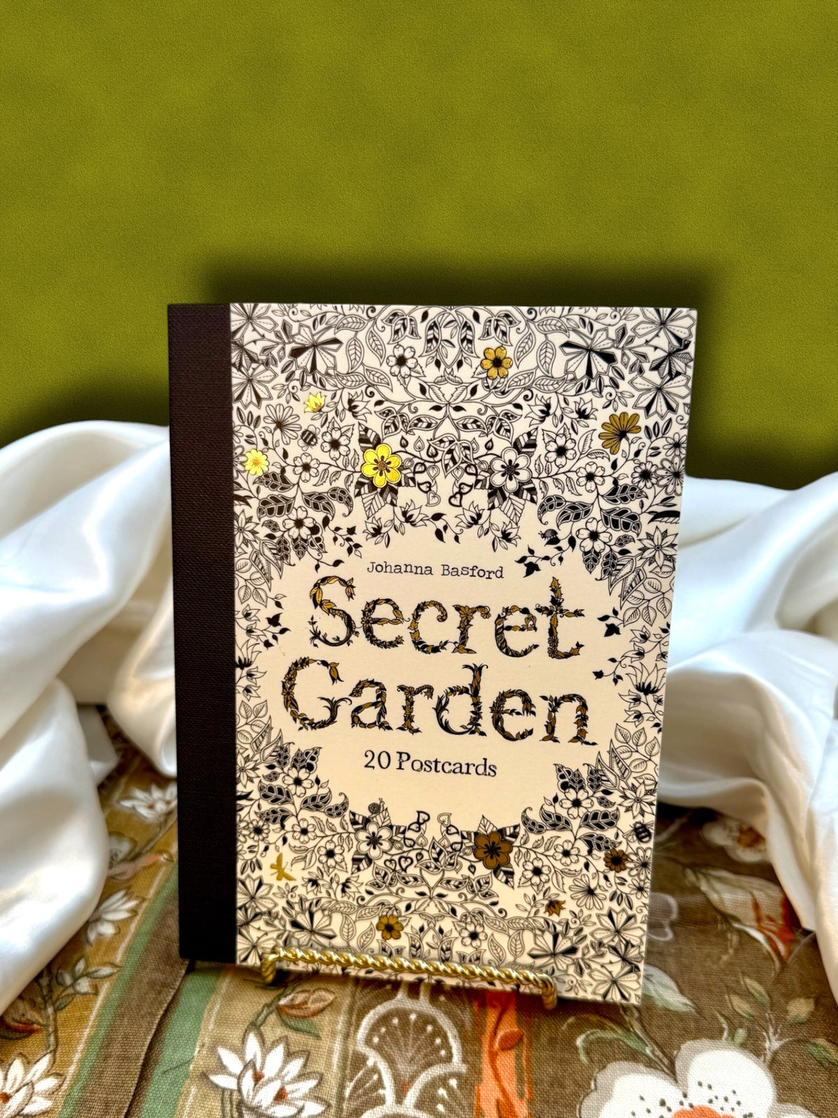 Secret Garden by Johanna Basford, collection of 20 detachable coloring postcards, displayed upright against an olive green backdrop with decorative fabric.