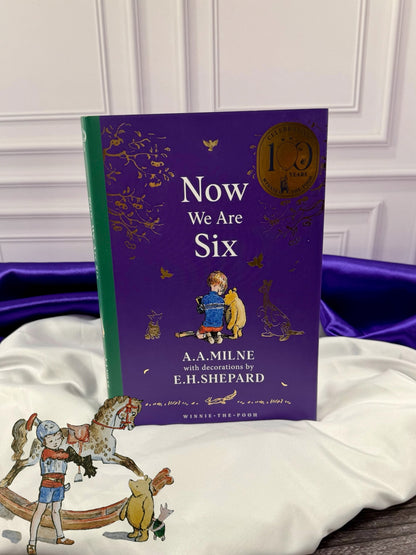 Now We Are Six by A. A. Milne, 100th anniversary hardcover with purple cover, green spine, gold anniversary emblem, and E. H. Shepard illustration of Christopher Robin with Winnie-the-Pooh.
