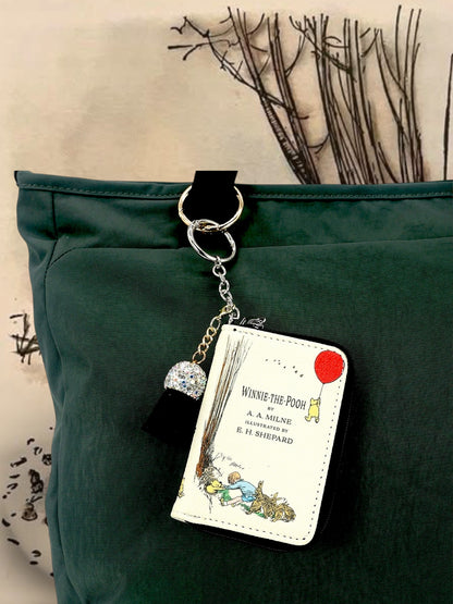 Winnie-the-Pooh miniature book bag charm hanging from a dark green handbag by its silver keyring, showing the illustrated cover and decorative tassel.