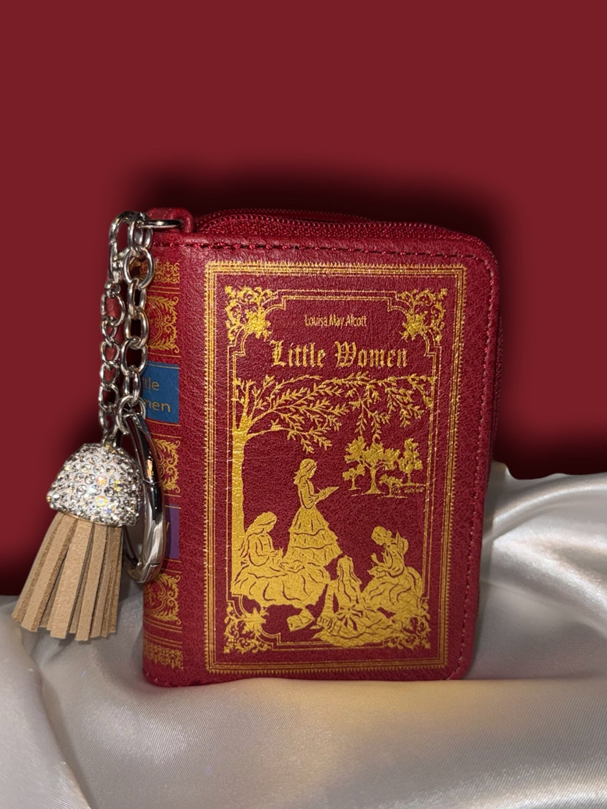 Little Women miniature book bag charm and coin purse in deep red with gold cover illustrations, silver keyring, and decorative tassel, displayed on ivory satin.