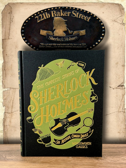Close-up of The Complete Stories of Sherlock Holmes clothbound hardcover showing gold lettering on a green illustrated design with violin, magnifying glass, pipe, deerstalker hat and detective motifs.