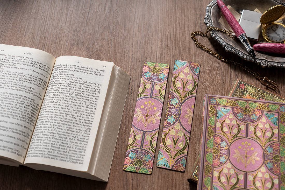 Three decorative paper bookmarks with a pink, gold, and green Art Nouveau tree design are arranged on a wooden table next to an open book, with a matching patterned book cover and stationery in the background.