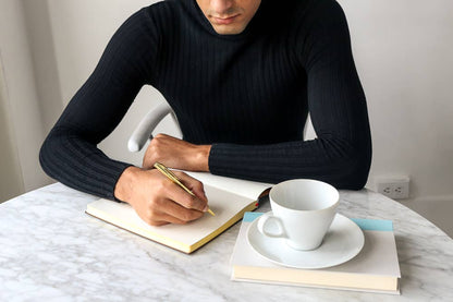 A man in a black ribbed sweater sits at a marble table, writing in an open notebook with a gold pen. A white coffee cup on a saucer and a stack of books are on the table in front of him.