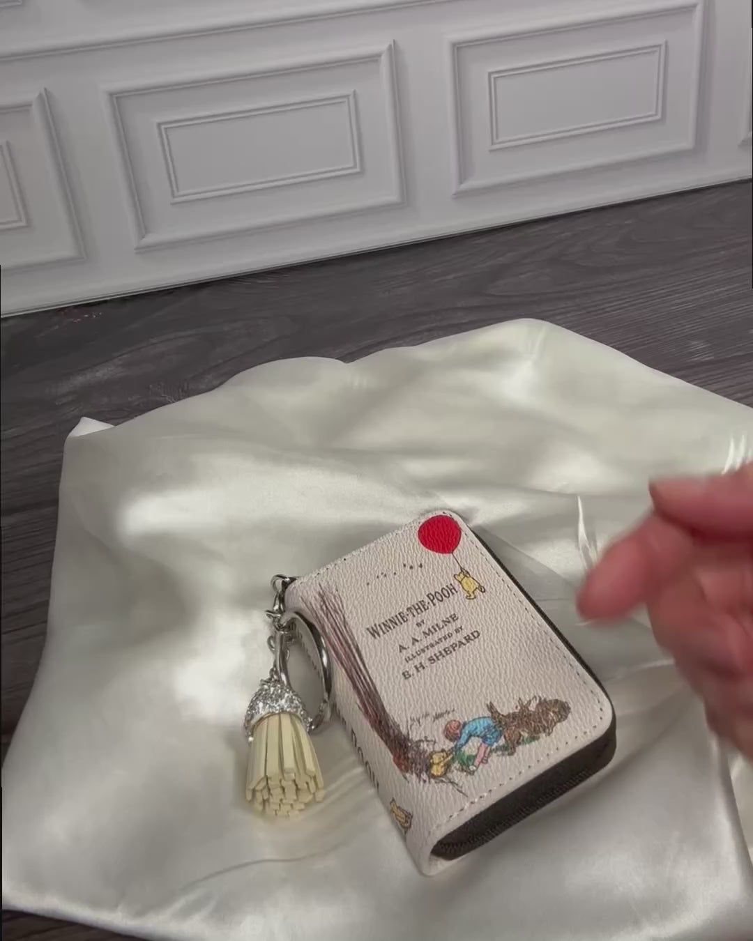 Winnie-the-Pooh miniature book coin purse and bag charm featuring E. H. Shepard illustrations, a zippered compartment, silver keyring, and decorative tassel. A charming literary accessory from StoryGiftStudio™.
